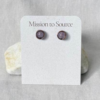 A pair of small round stud earrings with light purple stones in a copper wire wrap setting, displayed on a white card labeled "Mission to Source" against a light fabric background with two stones behind it.
