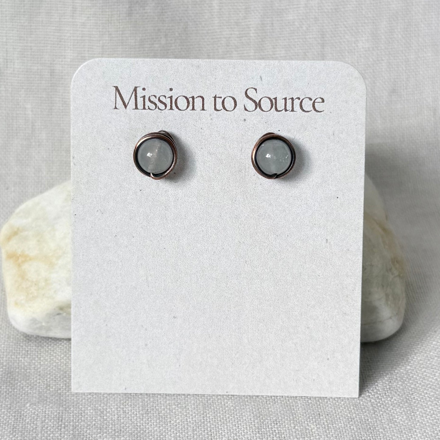 A pair of small wrap-style stud earrings with white round stones in copper wire settings, displayed on a white card that reads "Mission to Source," set against a light fabric background with two small stones.