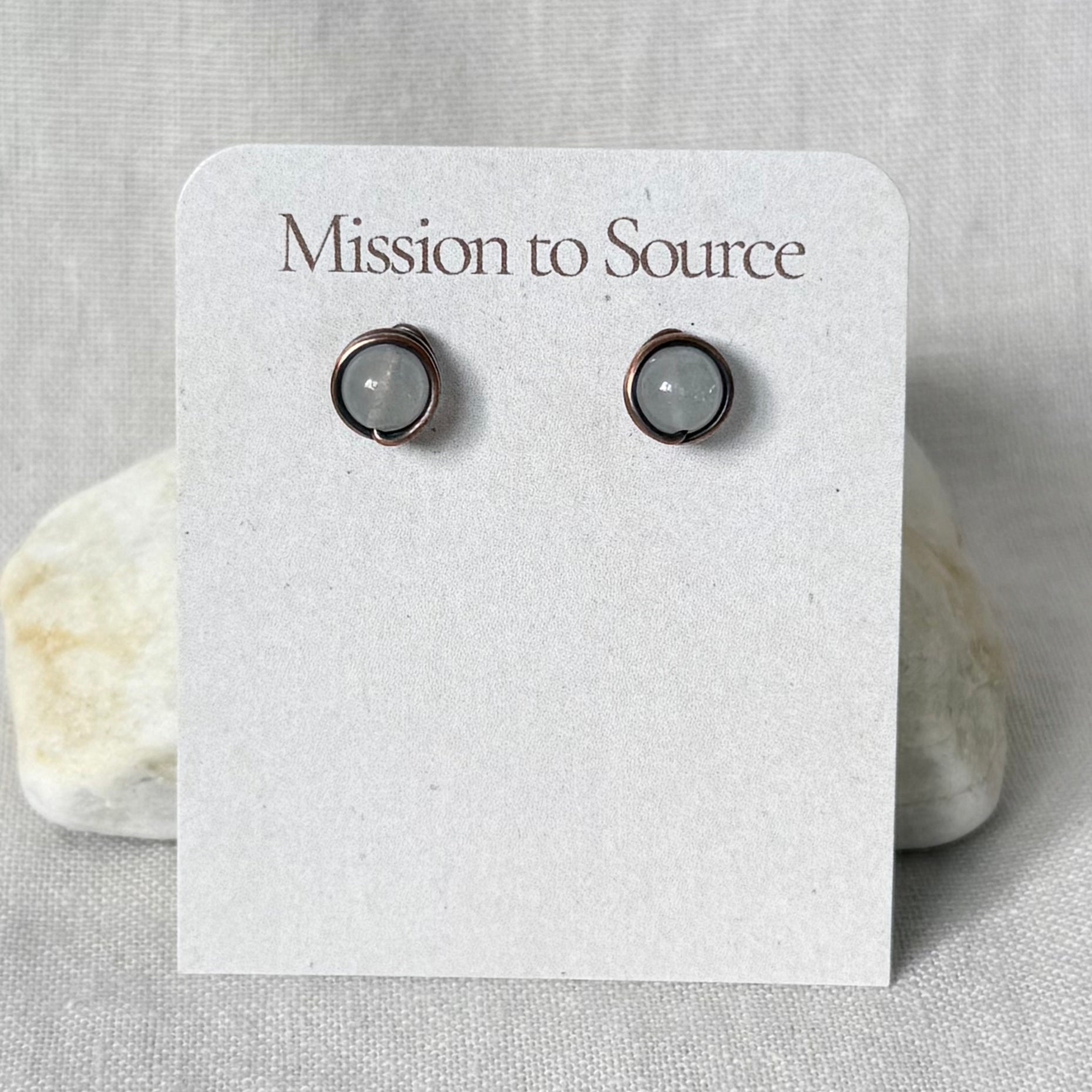 A pair of small wrap-style stud earrings with white round stones in copper wire settings, displayed on a white card that reads "Mission to Source," set against a light fabric background with two small stones.