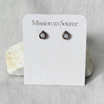 A pair of small round wire wrapped stud earrings, displayed on a white card labeled "Mission to Source" against a light fabric background with a stone prop.