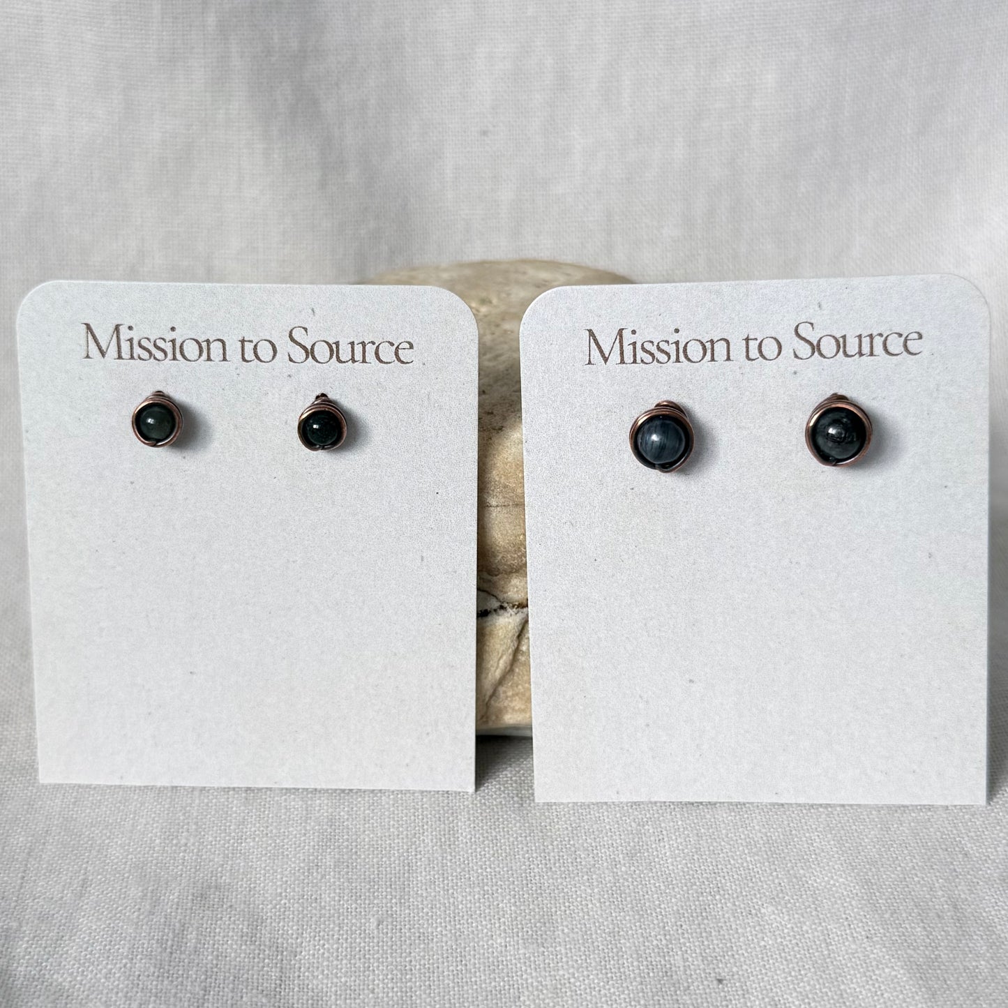 Two pairs of small stud earrings displayed on white card holders that read 'Mission to Source.' The earrings have dark round stones in warm copper wire settings, photographed against a light fabric backdrop.