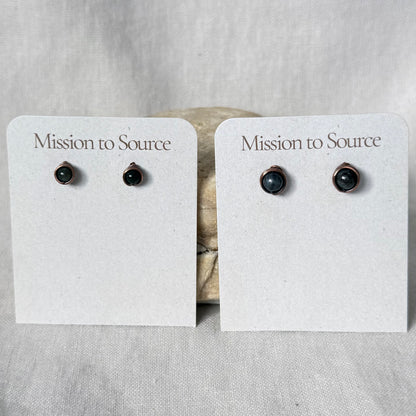 Two pairs of small stud earrings displayed on white card holders that read 'Mission to Source.' The earrings have dark round stones in warm copper wire settings, photographed against a light fabric backdrop.