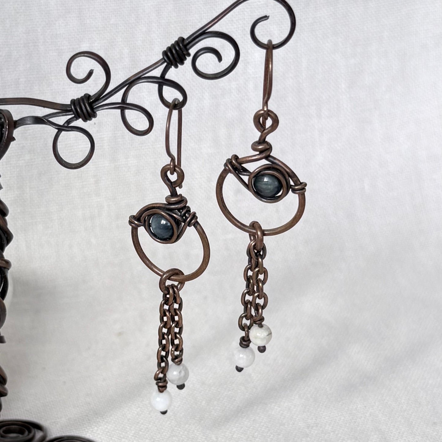 A pair of handmade dangle earrings in copper wire wrapping, featuring a dark blue Apatite stone and small white bead drops, displayed on a decorative wire stand against a light grey fabric background.