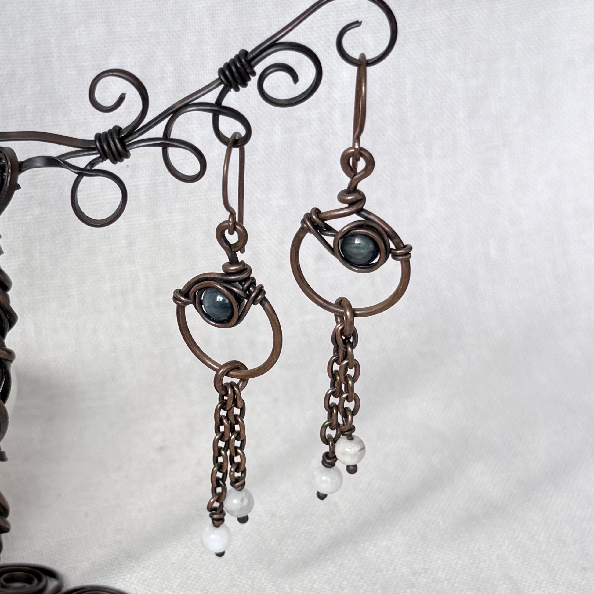 A pair of handmade dangle earrings in copper wire wrapping, featuring a dark blue Apatite stone and small white bead drops, displayed on a decorative wire stand against a light grey fabric background.