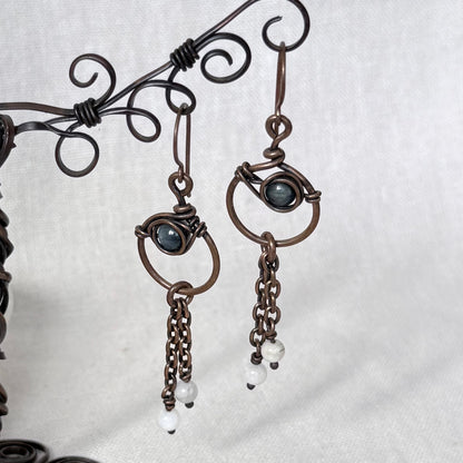 A pair of handmade dangle earrings in copper wire wrapping, featuring a dark blue Apatite stone and small white bead drops, displayed on a decorative wire stand against a light grey fabric background.