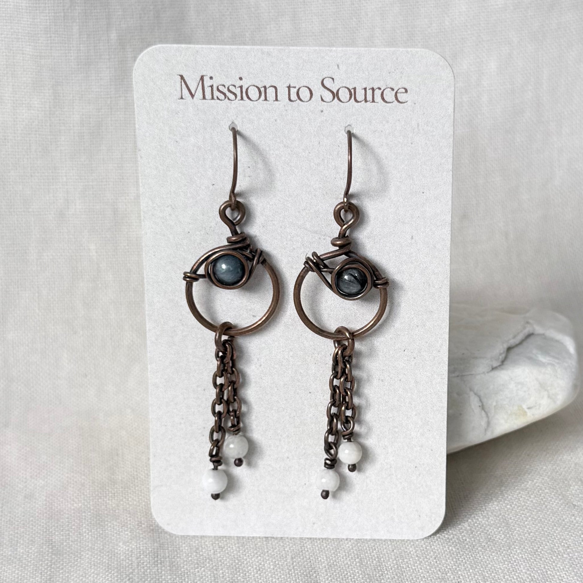 A pair of handmade copper dangle earrings, featuring small oval blue-grey stones and dangling chain elements, displayed on a white card labeled 'Mission to Source' against a neutral fabric background.