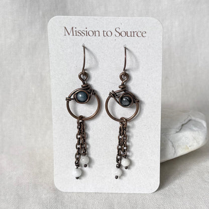 A pair of handmade copper dangle earrings, featuring small oval blue-grey stones and dangling chain elements, displayed on a white card labeled 'Mission to Source' against a neutral fabric background.