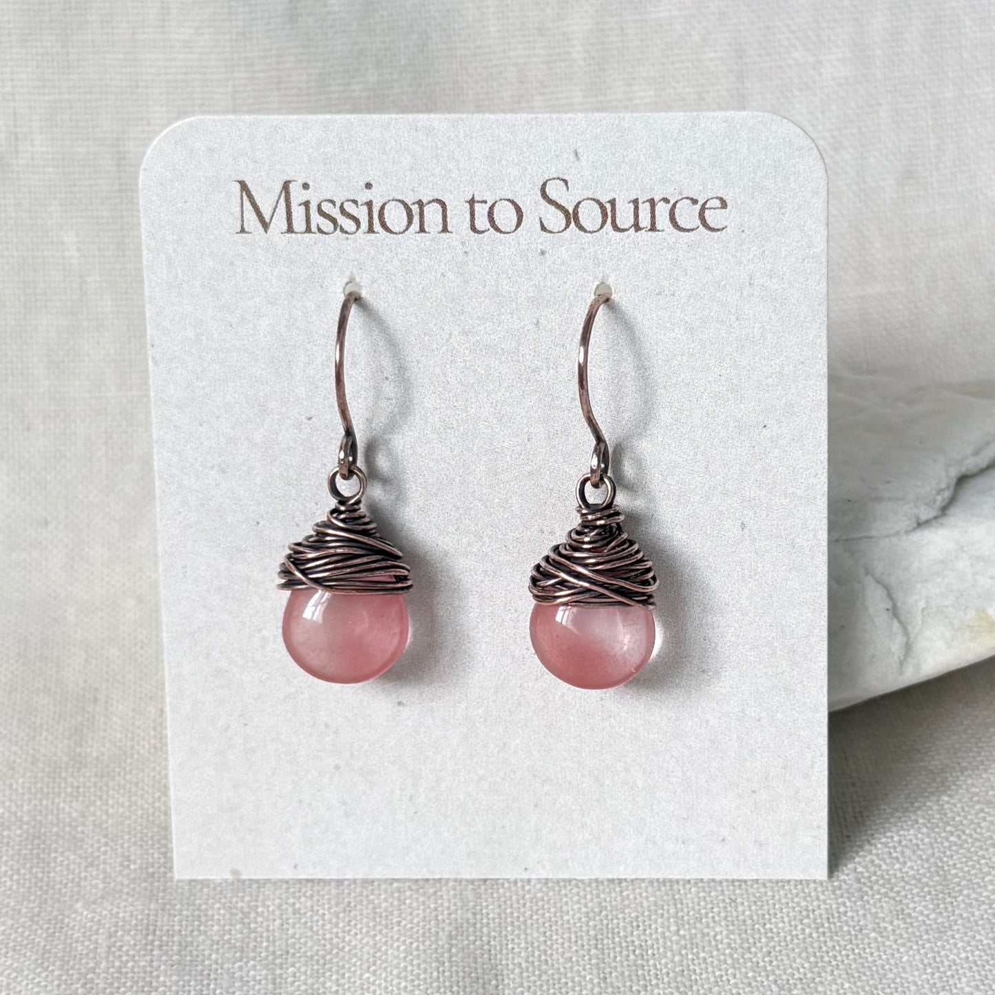 A pair of handmade dangle earrings with pink round beads wrapped in dark copper wire, displayed on a white card labeled 'Mission to Source'.
