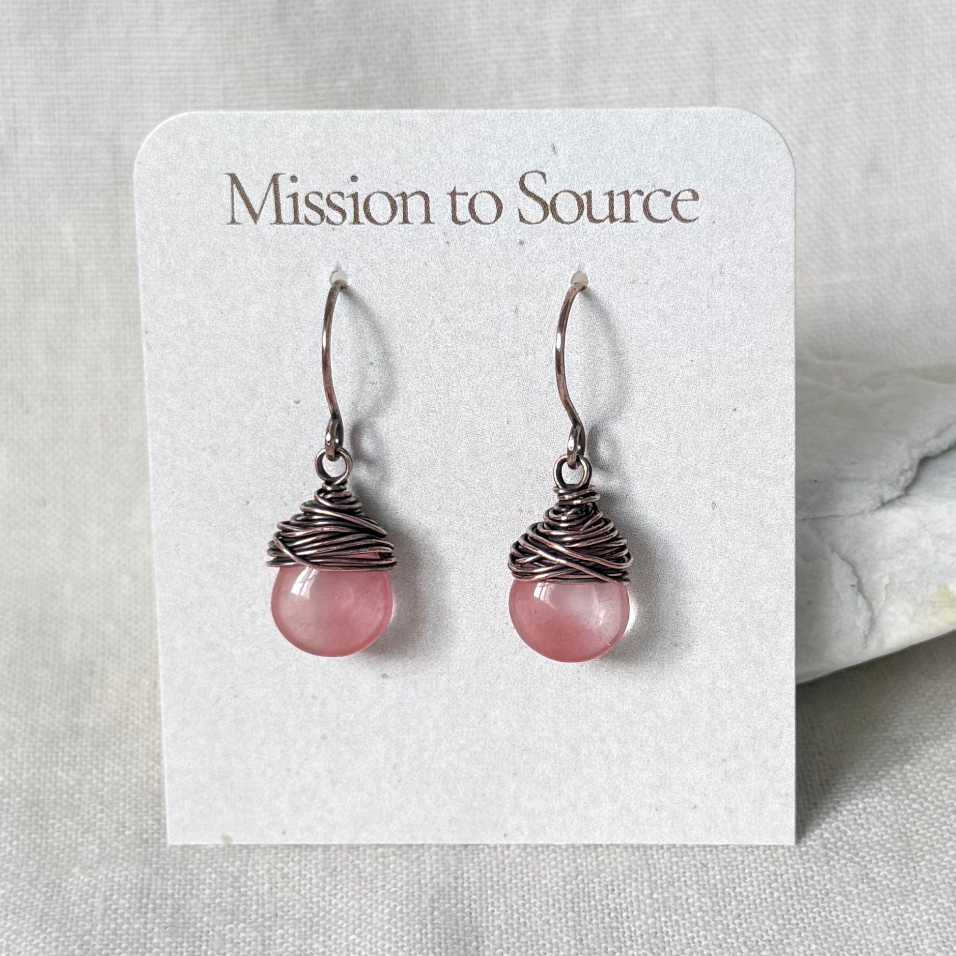 A pair of handmade dangle earrings with pink round beads wrapped in dark copper wire, displayed on a white card labeled 'Mission to Source'.