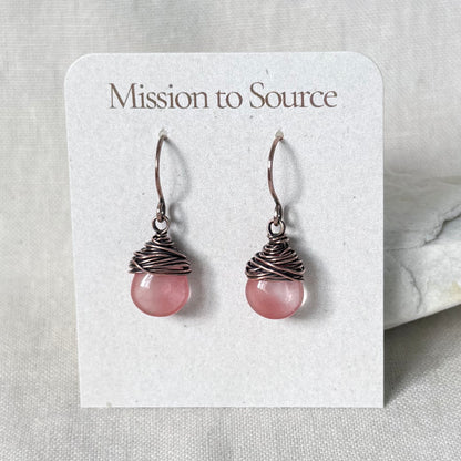 A pair of handmade dangle earrings with pink round beads wrapped in dark copper wire, displayed on a white card labeled 'Mission to Source'.