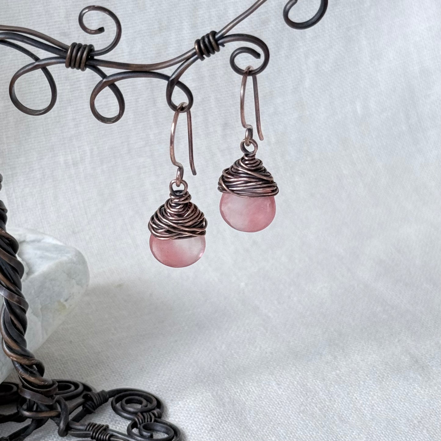 A pair of handmade dangle earrings with round pink stone drops wrapped in dark copper wire, hanging from a decorative dark metal wire stand against a white fabric background.