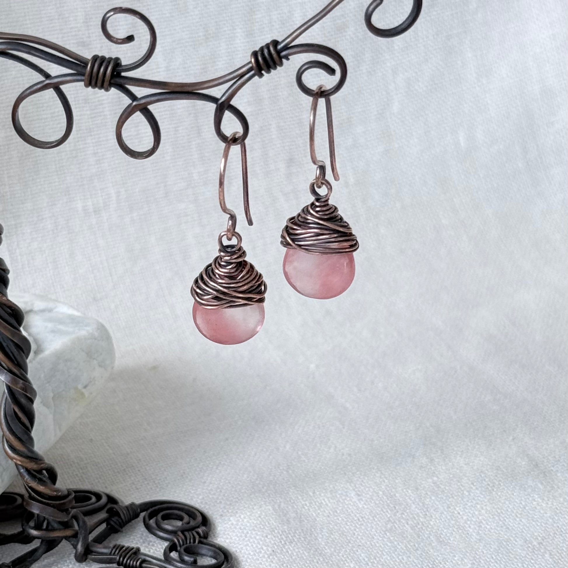 A pair of handmade dangle earrings with round pink stone drops wrapped in dark copper wire, hanging from a decorative dark metal wire stand against a white fabric background.