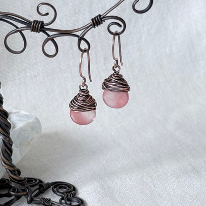 A pair of handmade dangle earrings with round pink stone drops wrapped in dark copper wire, hanging from a decorative dark metal wire stand against a white fabric background.