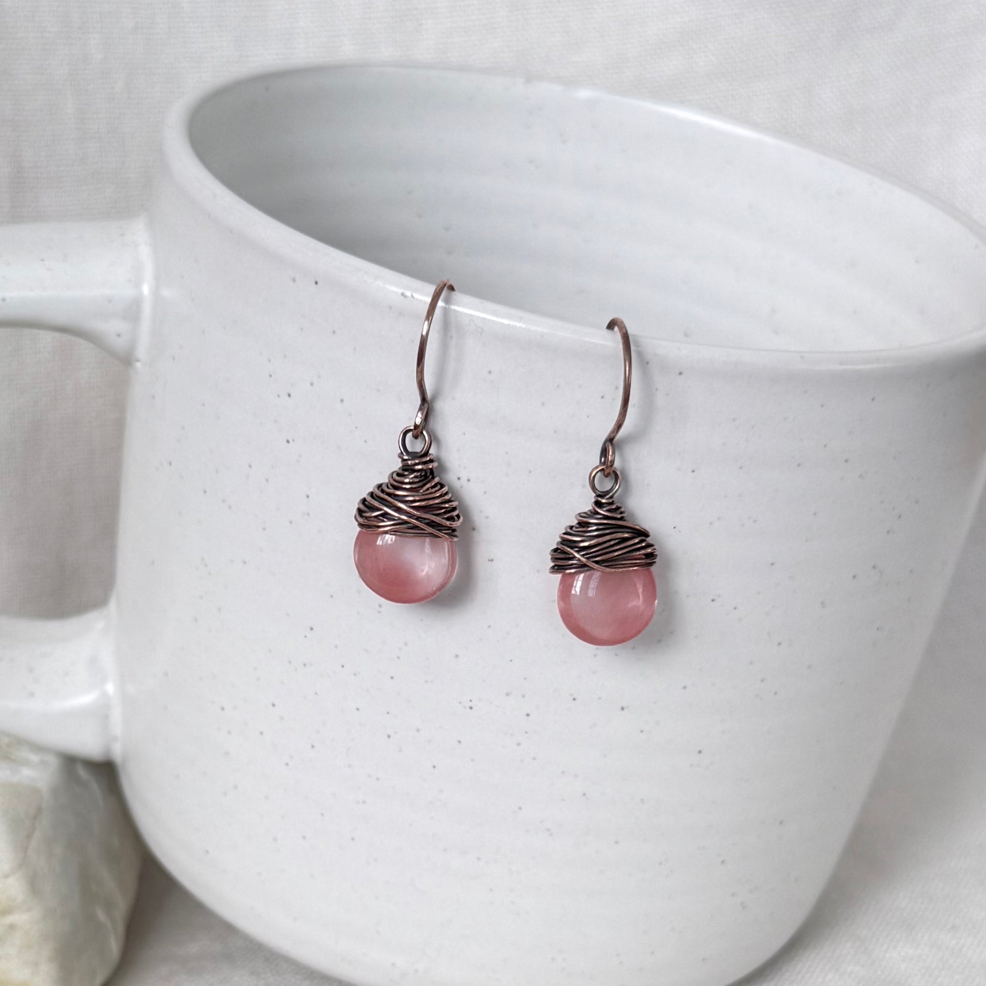 A pair of handmade dangle earrings with pink round beads wrapped in copper wire, resting against a white speckled ceramic mug.
