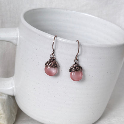 A pair of handmade dangle earrings with pink round beads wrapped in copper wire, resting against a white speckled ceramic mug.