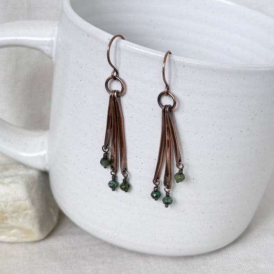 A pair of handmade copper drop earrings with multiple thin wire strands and small green bead charms, hanging from the rim of a white speckled ceramic mug.