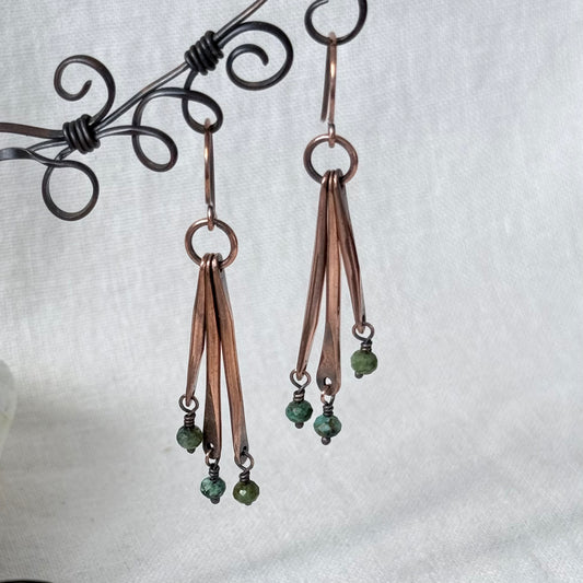 A pair of handmade dangle earrings with copper-toned metal strips and small green bead accents, displayed on a black wire stand against a light fabric background.