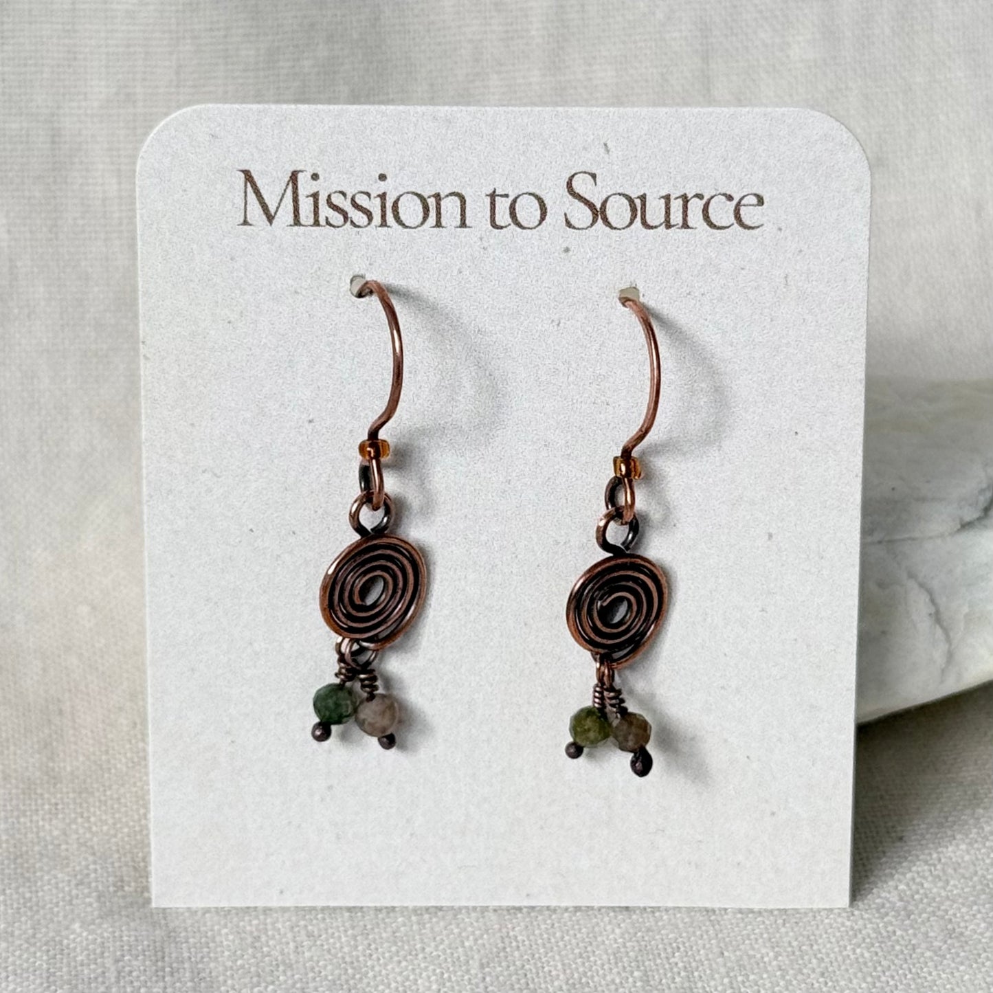 A pair of handmade copper wire hoop earrings with spiral details and small green and grey beads, displayed on a white card labeled 'Mission to Source'.