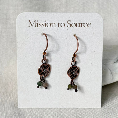 A pair of handmade copper wire hoop earrings with spiral details and small green and grey beads, displayed on a white card labeled 'Mission to Source'.