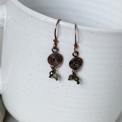 A pair of handmade copper wire hoop earrings with spiral coil charms and small green bead accents, hanging against a white speckled ceramic mug.