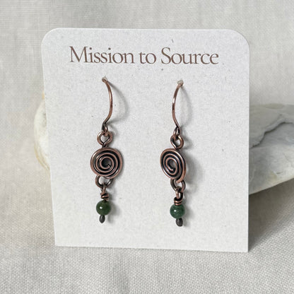 A pair of handmade dangle earrings displayed on a white card labeled 'Mission to Source.' The earrings have copper spiral wire details and small dark green beads at the bottom, set against a neutral fabric background.