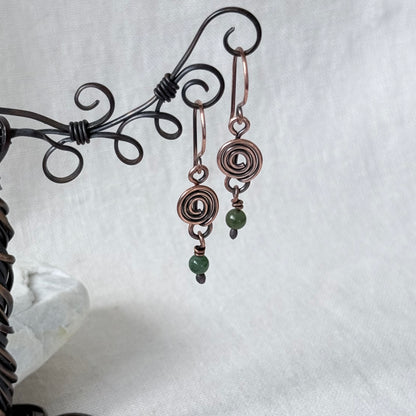 A pair of handmade copper wire drop earrings with green bead accents, displayed on a black metal stand against a white fabric background.