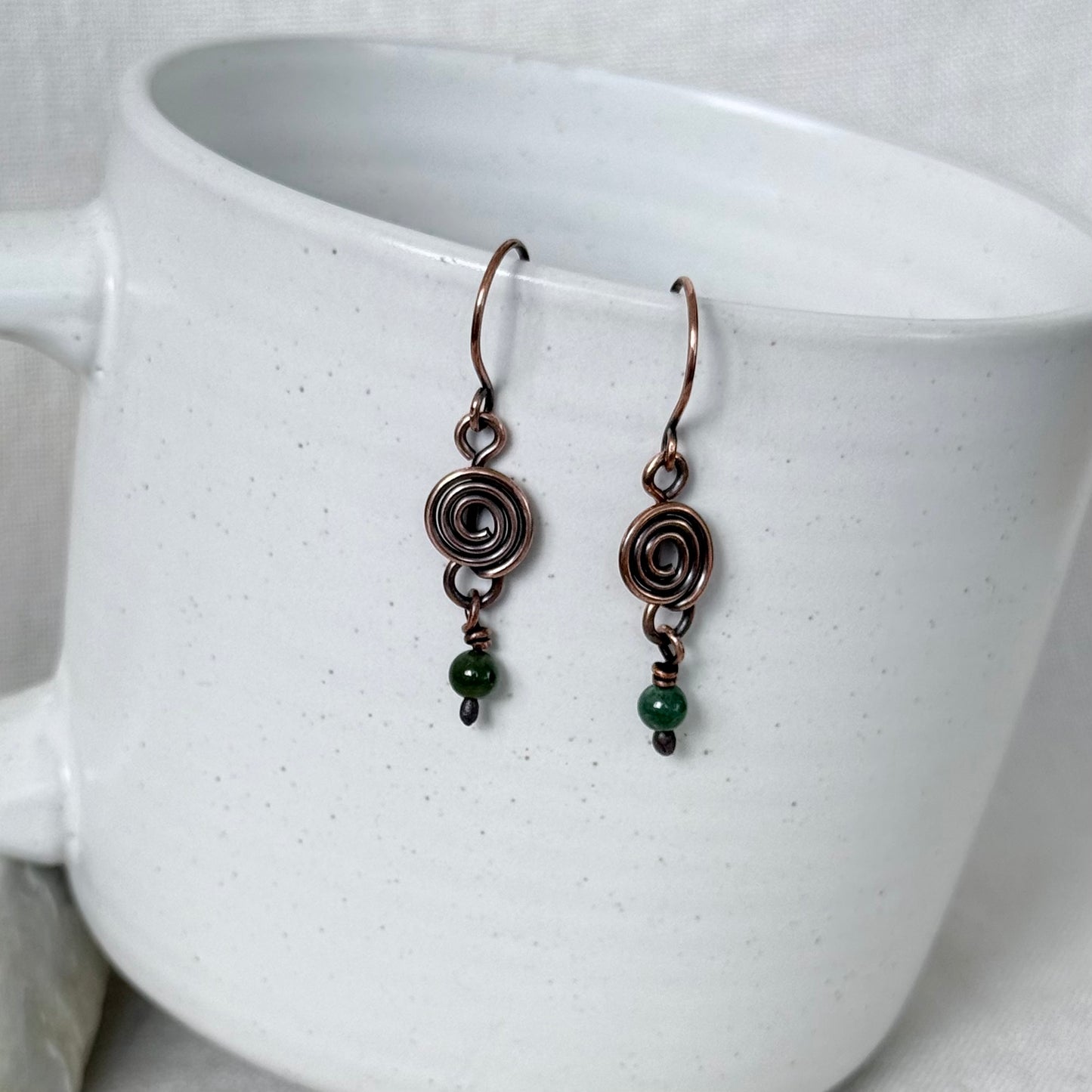 A pair of handmade copper drop earrings with spiral wire charms and small green beads, hanging from the rim of a speckled white ceramic mug.