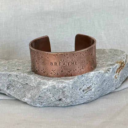 Copper cuff with 'BREATHE' engraving on a stone surface and linen background