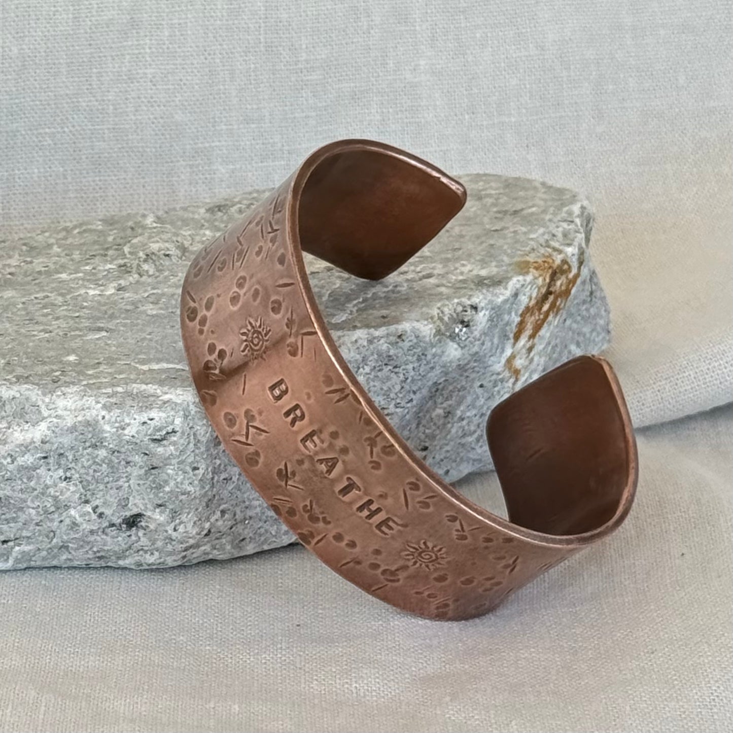 Copper cuff with 'BREATHE' engraving on a stone surface and linen background