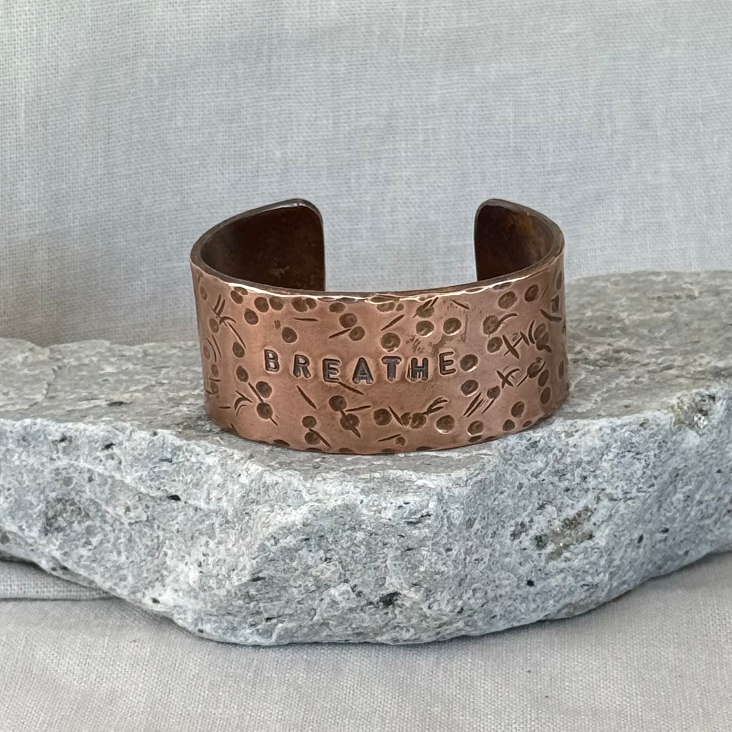 Copper cuff with 'BREATHE' engraving on a stone surface and linen background