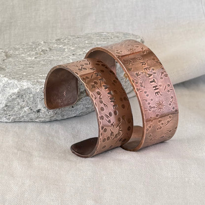 Two Copper cuffs with 'BREATHE' engraving on a stone surface and linen background