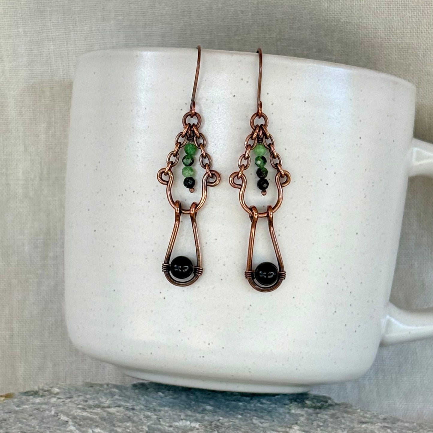 Copper earrings with green and black beads on a white mug with linen background