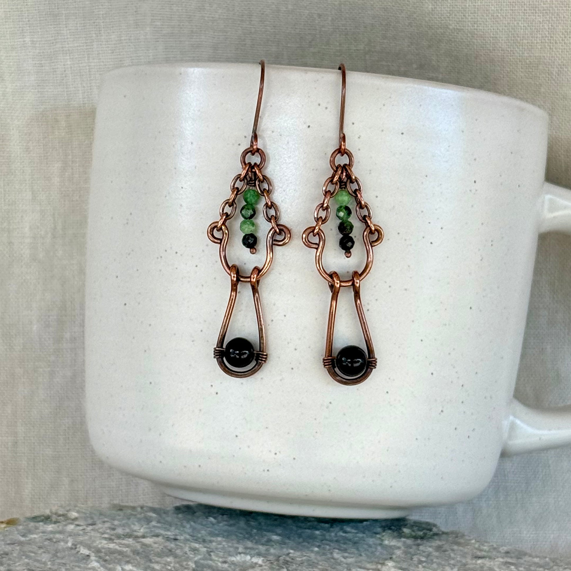 Copper earrings with green and black beads on a white mug with linen background