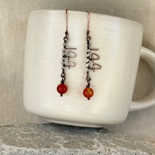 Pair of copper wire earrings forming the number 144 with orange beads on a white mug against linen background