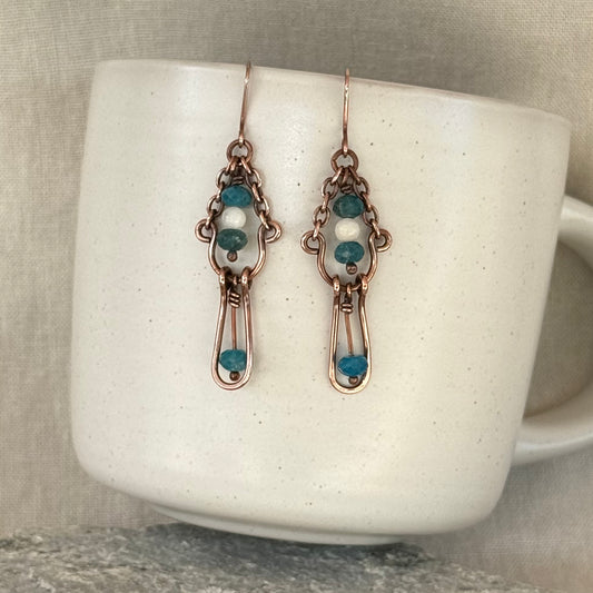 a pair of copper earrings with blue and white stones hanging from a bright cream mug on stone surface with linen background