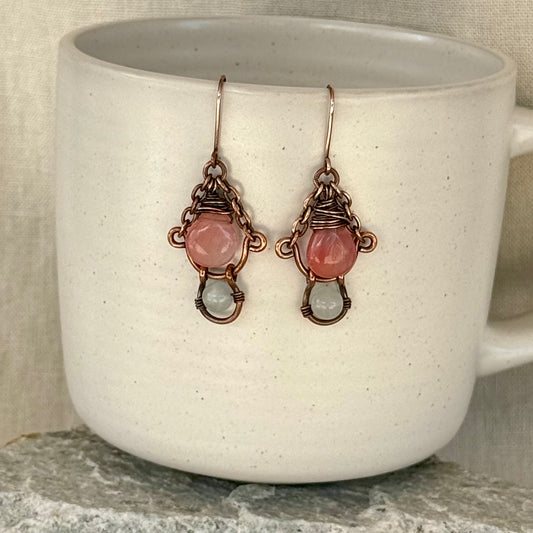 pair of copper earrings with pink and white beads hanging from a mug with stone surface and linen background