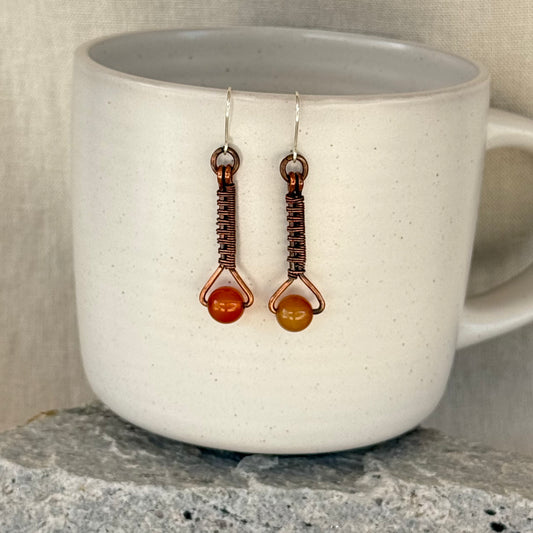 Pair of copper woven earrings with carnelian bead and silver earhooks hanging from a white mug on stone surface with linen background