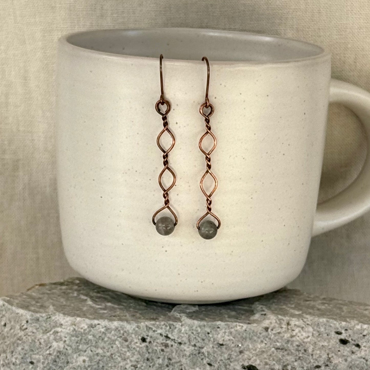 Copper earrings with beads hanging over a white mug on a stone surface.