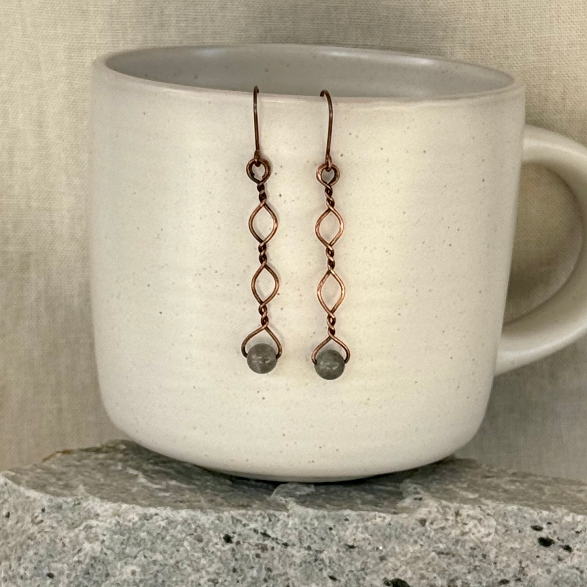 Copper earrings with beads hanging over a white mug on a stone surface.