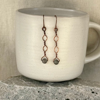 Copper earrings with beads hanging over a white mug on a stone surface.