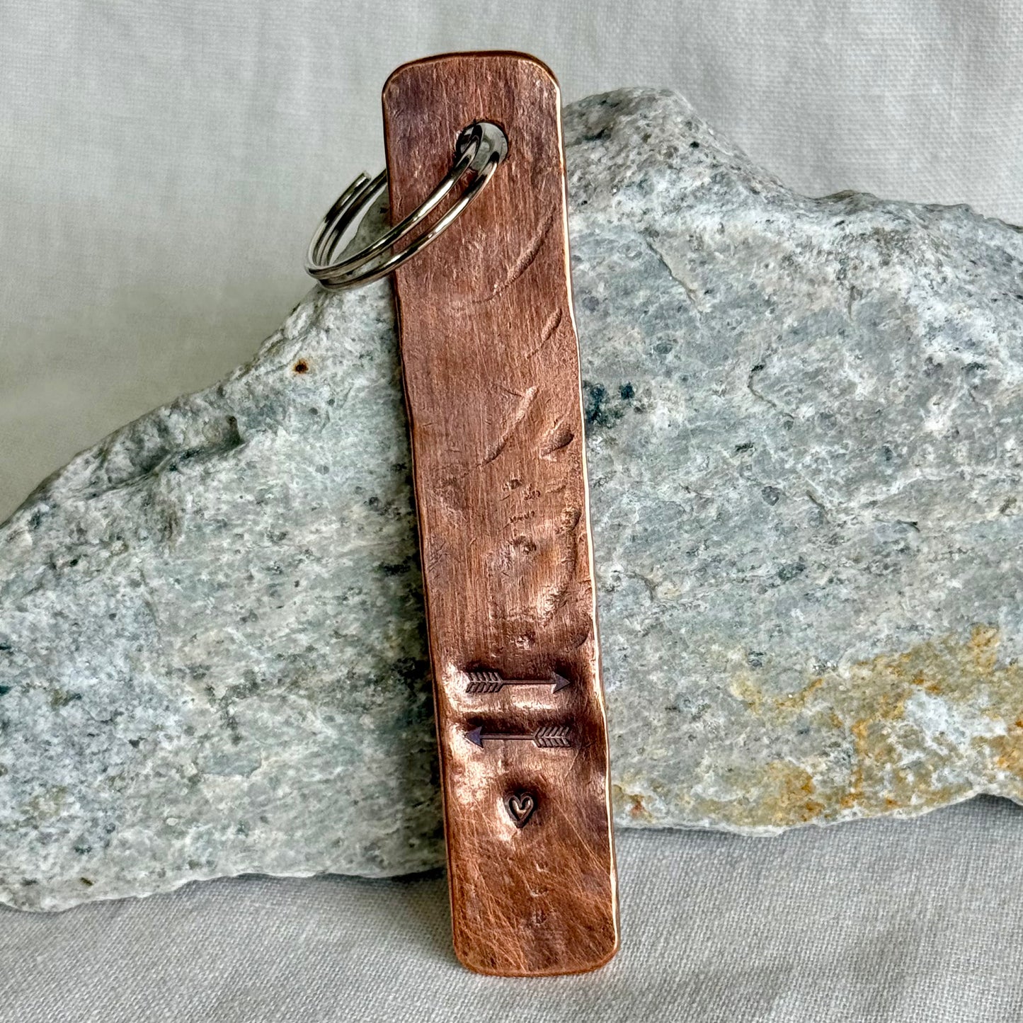 Copper keychain with two arrows and a heart stamped on it against a stone and linen background