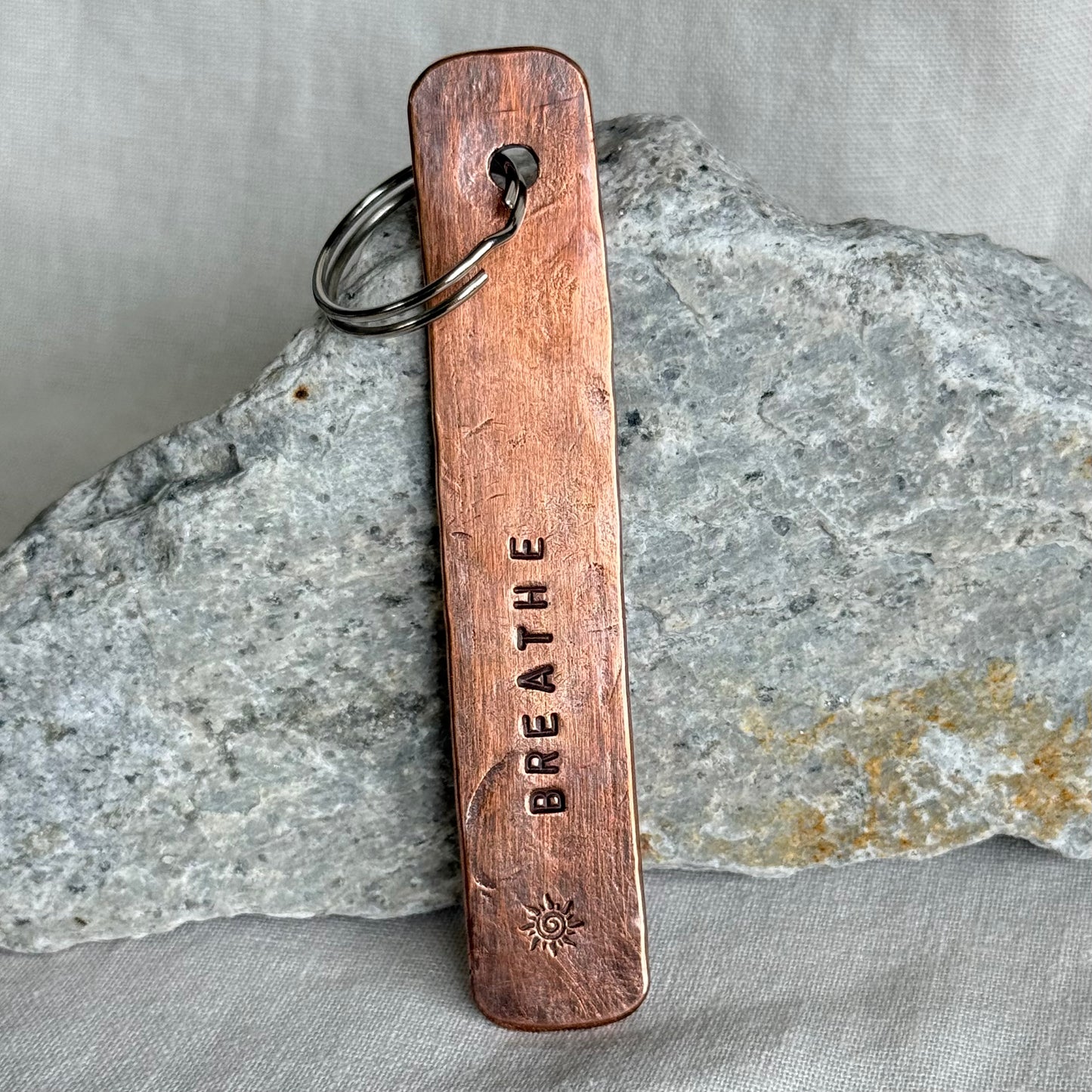Copper keychain with 'BREATHE' stamped on it against a stone and linen background