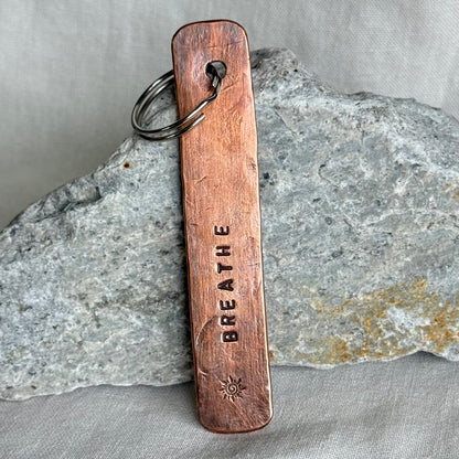 Copper keychain with 'BREATHE' stamped on it against a stone and linen background
