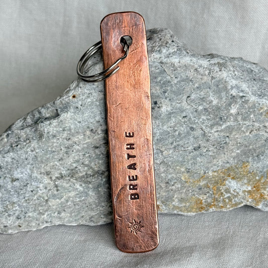 Copper keychain with 'BREATHE' stamped on it against a stone and linen background