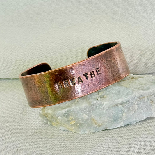 Copper cuff with 'BREATHE' engraving on a stone surface and linen background