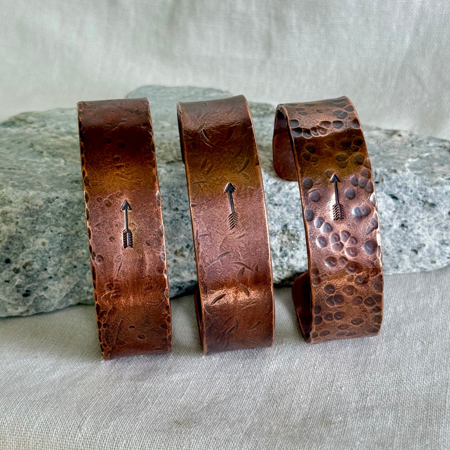 Three copper bracelets with stamped arrow designs on a stone and linen surface