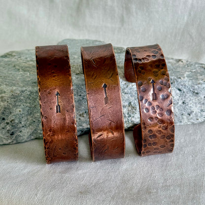 Three copper bracelets with stamped arrow designs on a stone and linen surface
