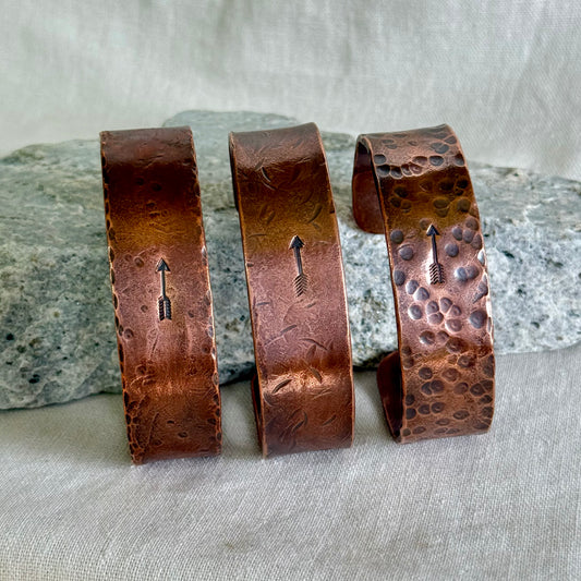 Three copper bracelets with stamped arrow designs on a stone and linen surface