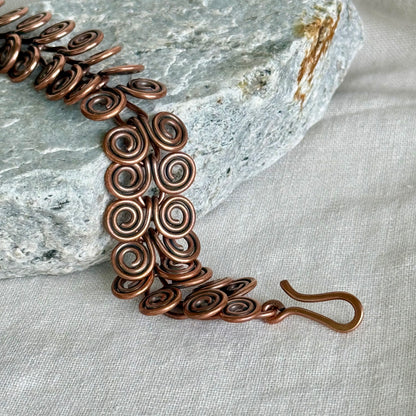 Copper bracelet with intricate spiral design on a stone and fabric background