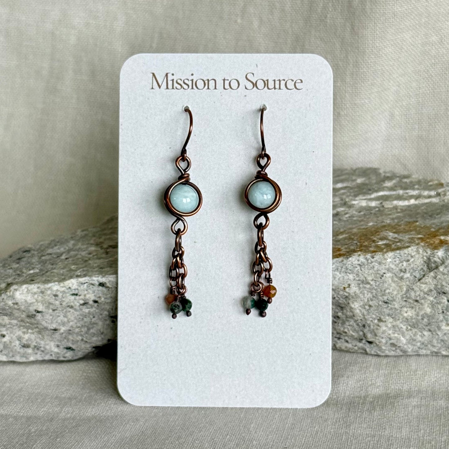 Pair of copper earrings on a 'Mission to Source' card with a stone background